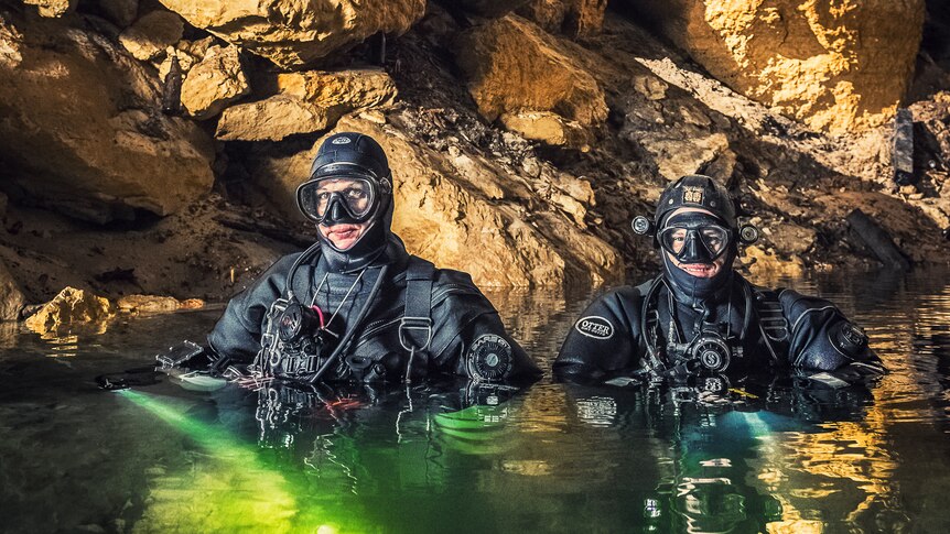 Two people wearing diving suits inside water inside a cave. 