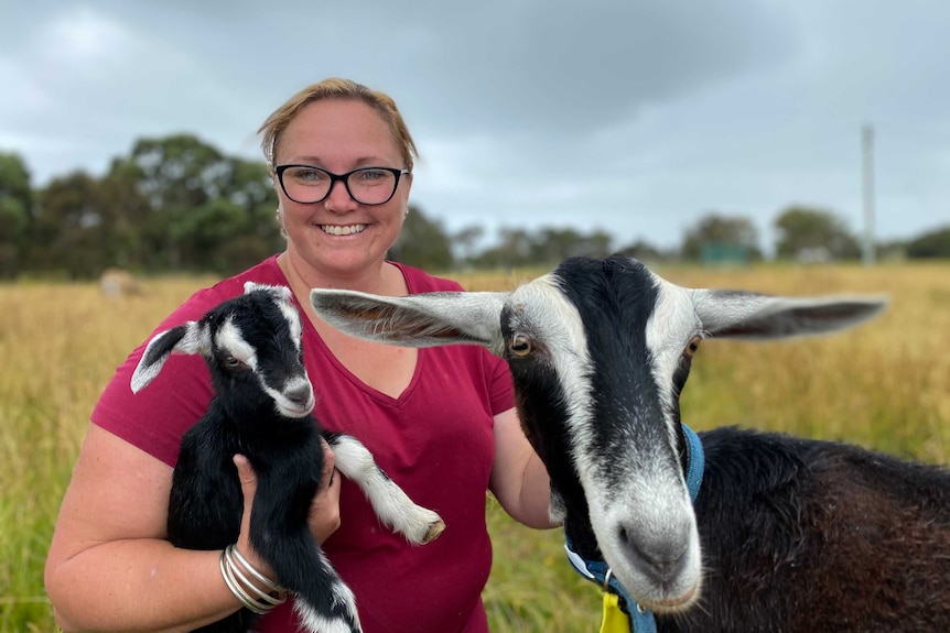 Alyssia Coates holds a kid and sits next to a larger goat.