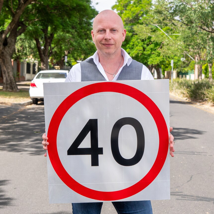 bald man holds 40km street sign in front of hime in middle of suburban street