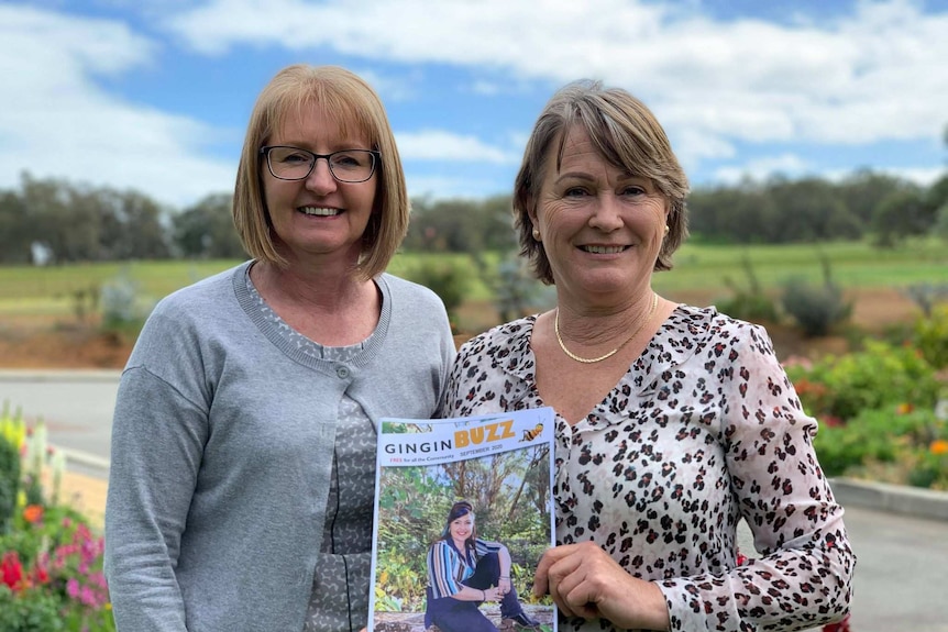 Two women in twinsets stand in front of a landscaped garden holding up a newsletter titled 'Gingin Buzz'.