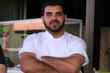 A chef looks directly into the camera with a solemn expression on his face and his arms crossed
