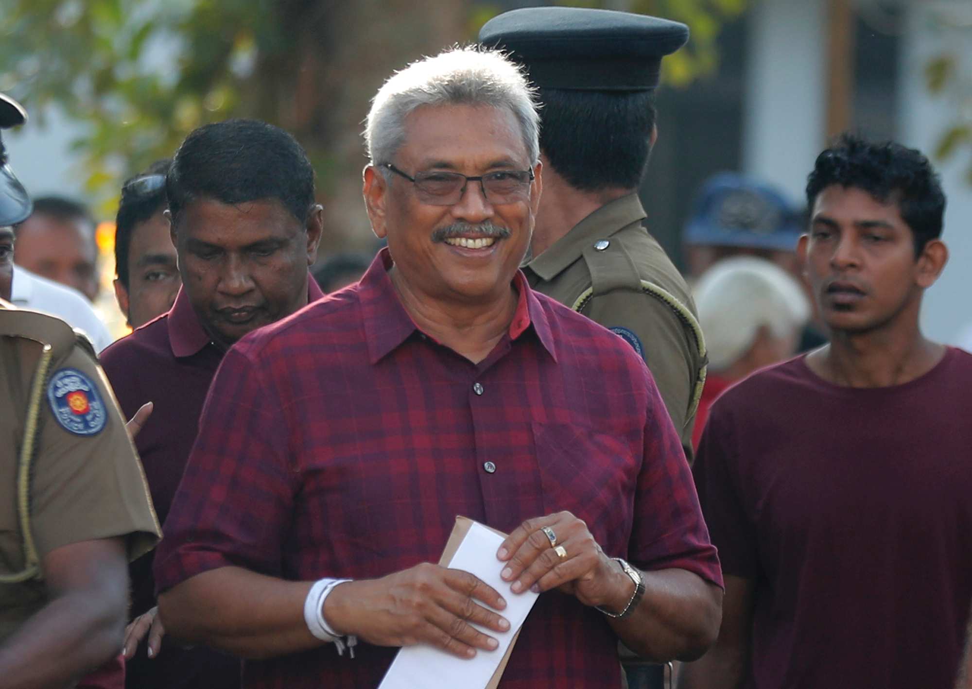 Smiling Rajapaksa, wearing glasses and a printed shirt, stands surrounded by a crowd of policemen and others.