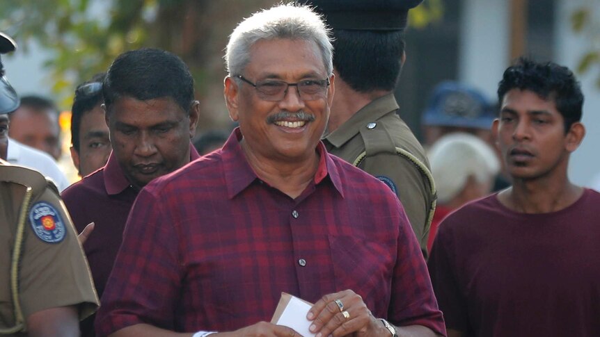 Smiling Rajapaksa, wearing glasses and a printed shirt, stands surrounded by a crowd of policemen and others.