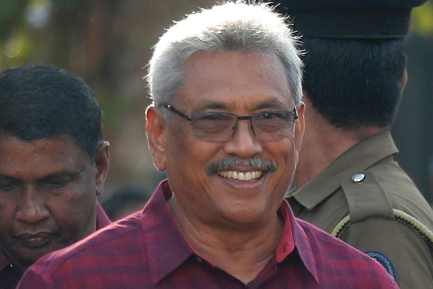 Smiling Rajapaksa, wearing glasses and a printed shirt, stands surrounded by a crowd of policemen and others.