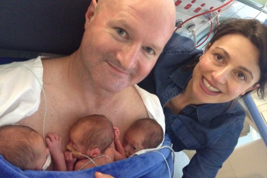 Scott Crank cradles his triplet sons with wife Lola. They are both smiling at the camera