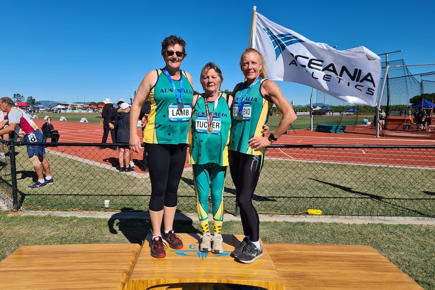 Three athletes in green and gold stand on a podium with a field in the background and a flag