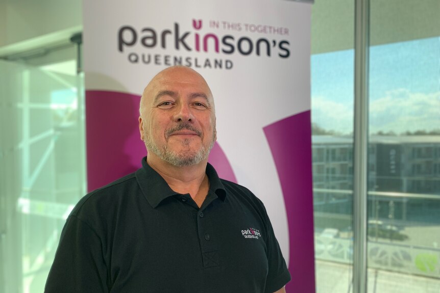 Parkinson’s Queensland CEO Miguel Diaz stands smiling in front of a sign at his office.