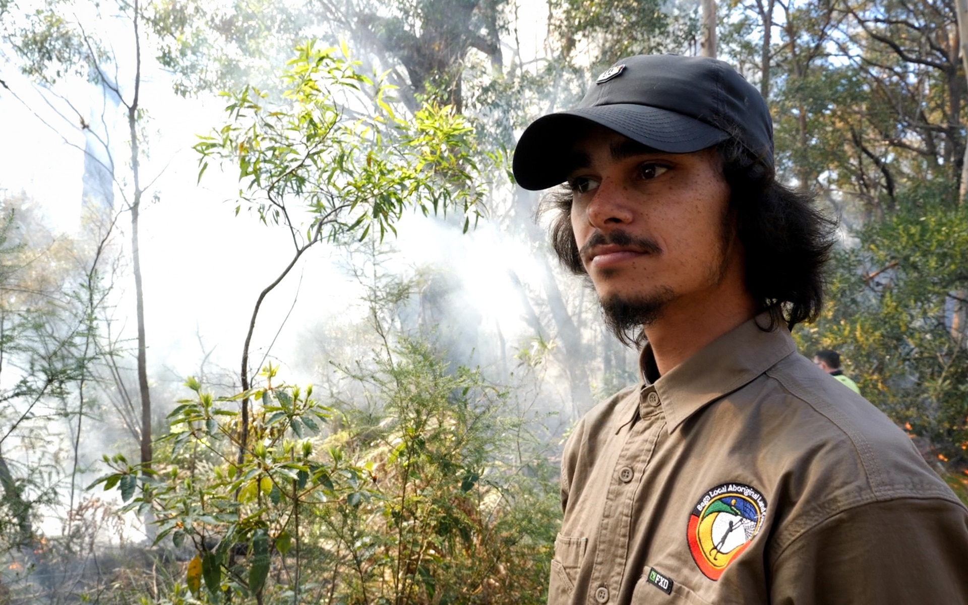 Un hombre con uniforme de guardabosques mira a lo lejos mientras el humo se desplaza por el bosque detrás de él.