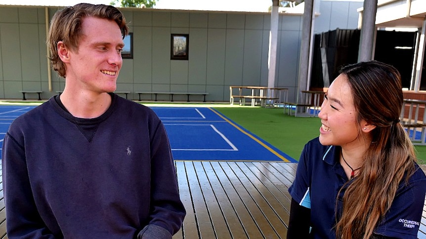 A man and a woman sitting on a bench in a school yard, smile at each oither.