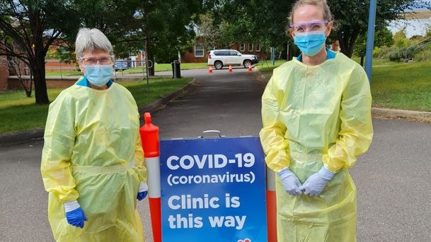 Two women wearing blue face masks and yellow plastic gowns stand beside a COVID-19 clinic sign.