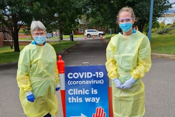 Two women wearing blue face masks and yellow plastic gowns stand beside a COVID-19 clinic sign.