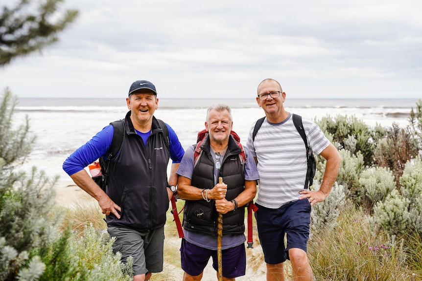 Three men in their 50s wearing walking gear and backpacks stand on the shore of a peaceful beach, smiling.
