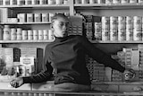 Young boy stars at camera from behind shop counter, with shelves full of tinned food behind him. Black and white photograph.