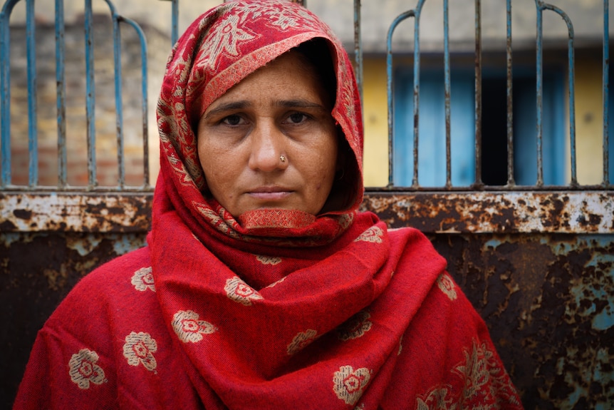 A woman wearing a red head scarf, with a gold nose stud, stares straight into camera while standing in front of a rusty fence 