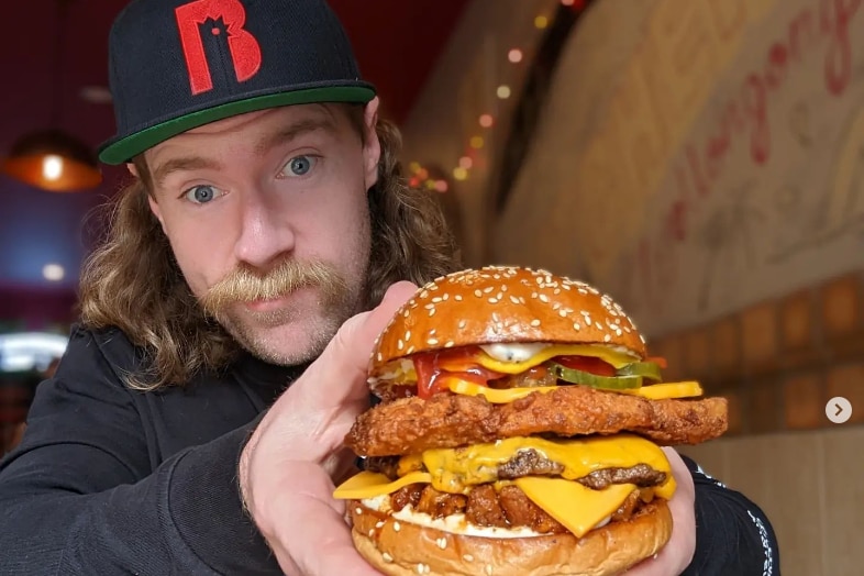 Man with moustache wearing a black and red cap holds big burger up to camera