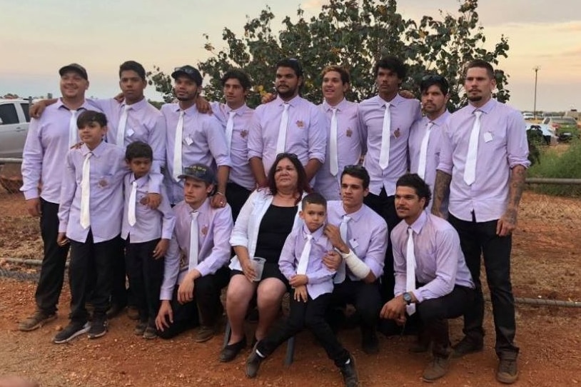 A woman sits outside in dusk light surrounded by a dozen young men dressed shirts and ties