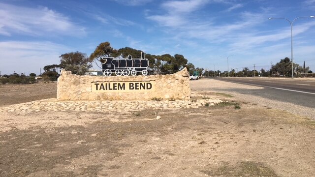 A sand coloured brick town sign has black letters reading 'Tailem Bend' and a black steam train. It is on a country road