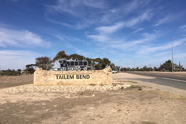 A sand coloured brick town sign has black letters reading 'Tailem Bend' and a black steam train. It is on a country road