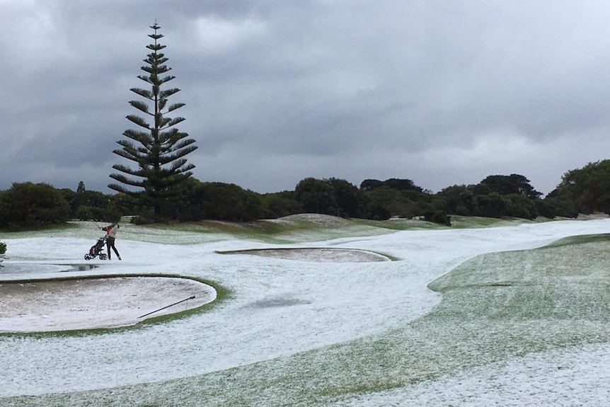 A person swings golf club on white golf course with snow over grass as ominous grey clouds are in sky.