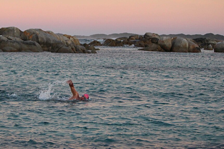 A man wearing a pink swimming cap and goggle swims in the ocean surrounded by granite boulders.