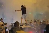 A man stands on a car in the middle of a smoky crowd at night.