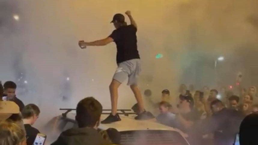 A man stands on a car in the middle of a smoky crowd at night.