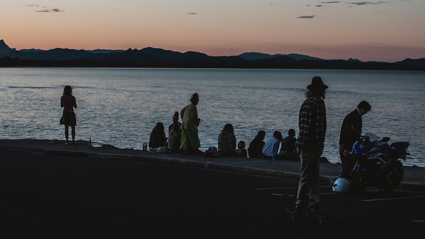 A group of people sit by the coastline at either sunset or sunrise.