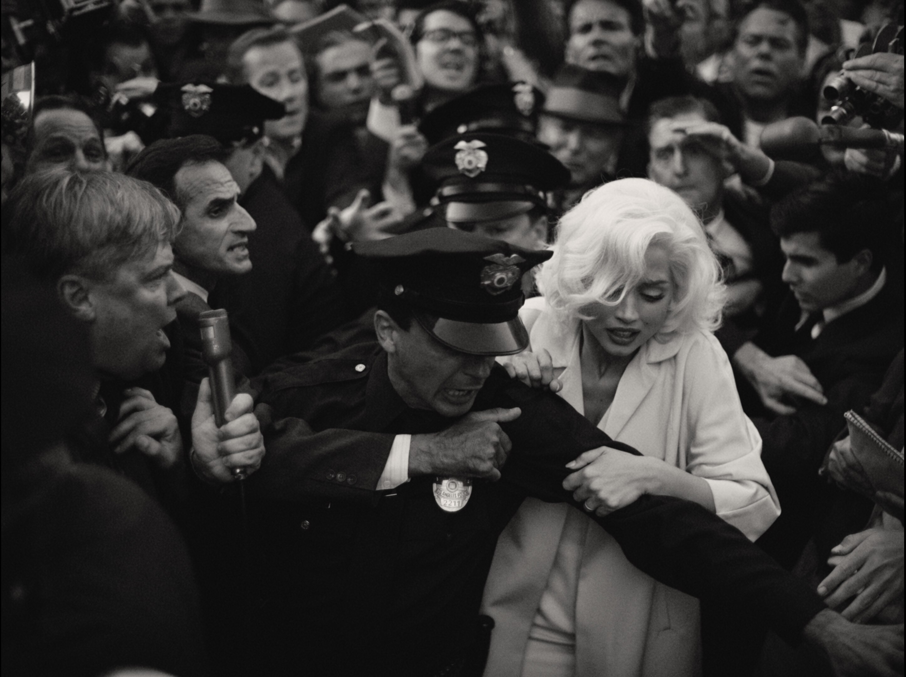 Black and white image of blonde white woman in white coat trying to squeeze through crowd of paparazzi.