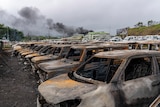 Dozens of torched cars sit in a carpark as smoke rises in the distance