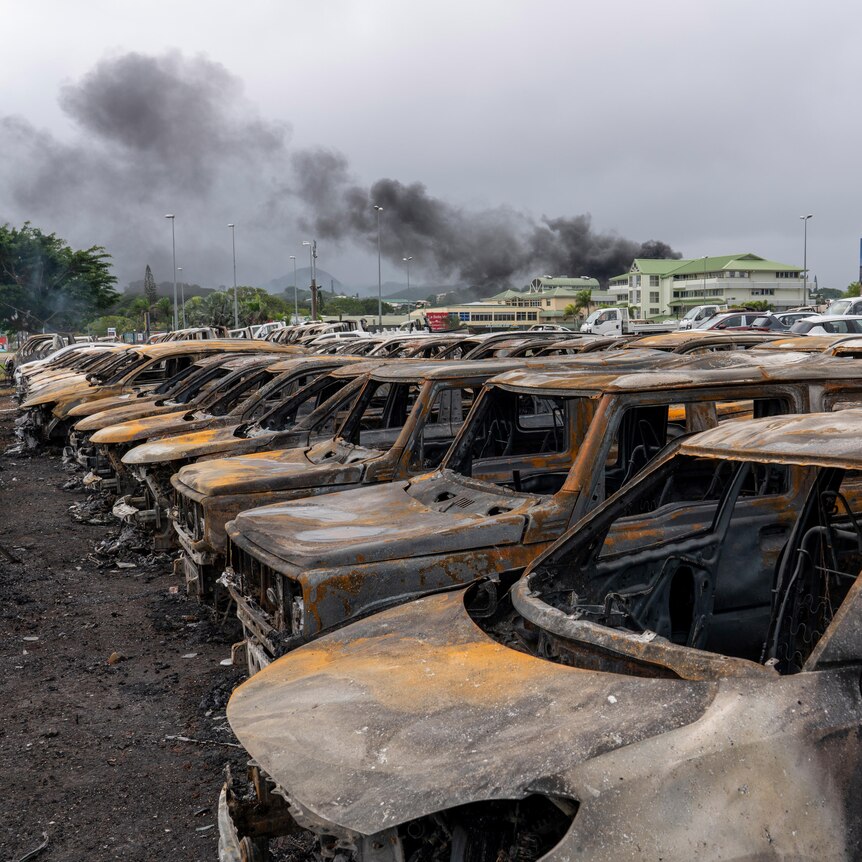 Dozens of torched cars sit in a carpark as smoke rises in the distance