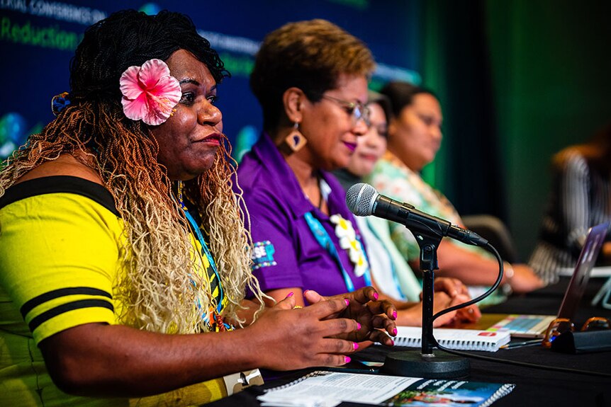 Women wearing yellow top and flower in ear speaks into mic sitting on panel table with other Pacific women. 