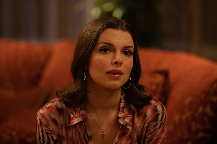 A woman with shoulder length brown hairs wears patterned shirt and hoop earrings sits on orange couch in warmly lit room.