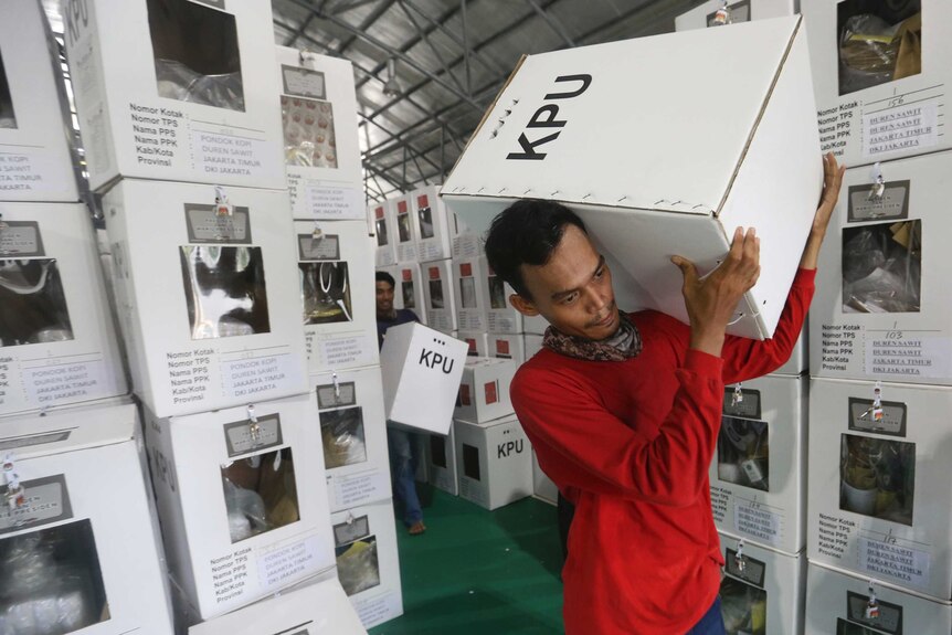 A man in a red shirt is carrying a large white ballot box marked "KPU" on his shoulder, surrounded by ballot boxes.