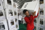 A man in a red shirt is carrying a large white ballot box marked "KPU" on his shoulder, surrounded by ballot boxes.