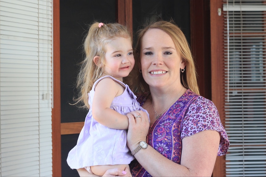 A mother in a purple top smiling at the camera holding her young daughter.