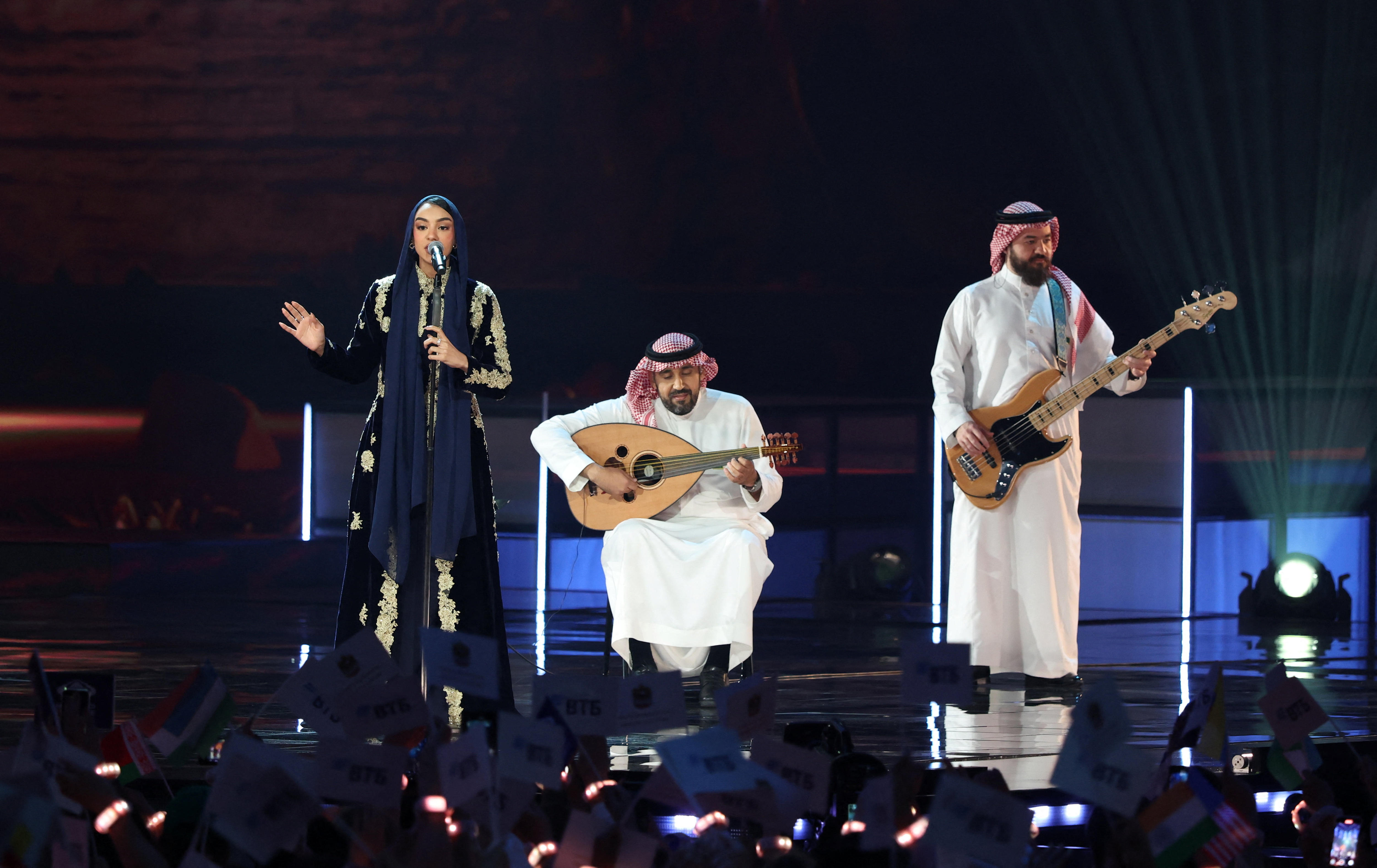 A woman wearing a black dress and headscarf sings near two men in white robes playing guitars