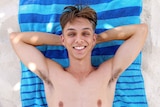 Connor Phillips smiles as he lies on a beach on a blue towel.