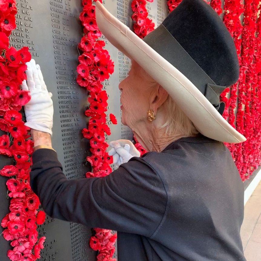 A finely dressed woman runs her fingers along a name engraved in stone, adorned with poppies.