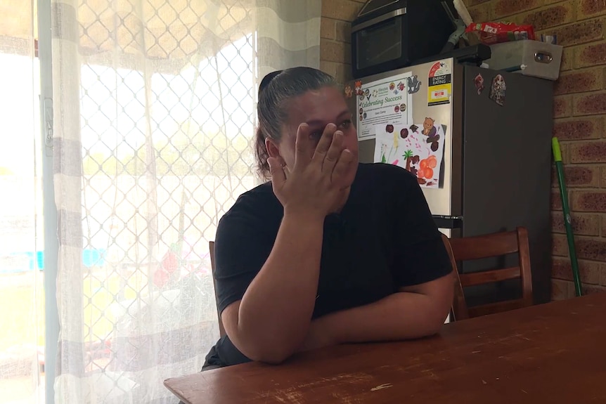 A woman in a black shirt sits at a kitchen table and wipes tears from her eyes.