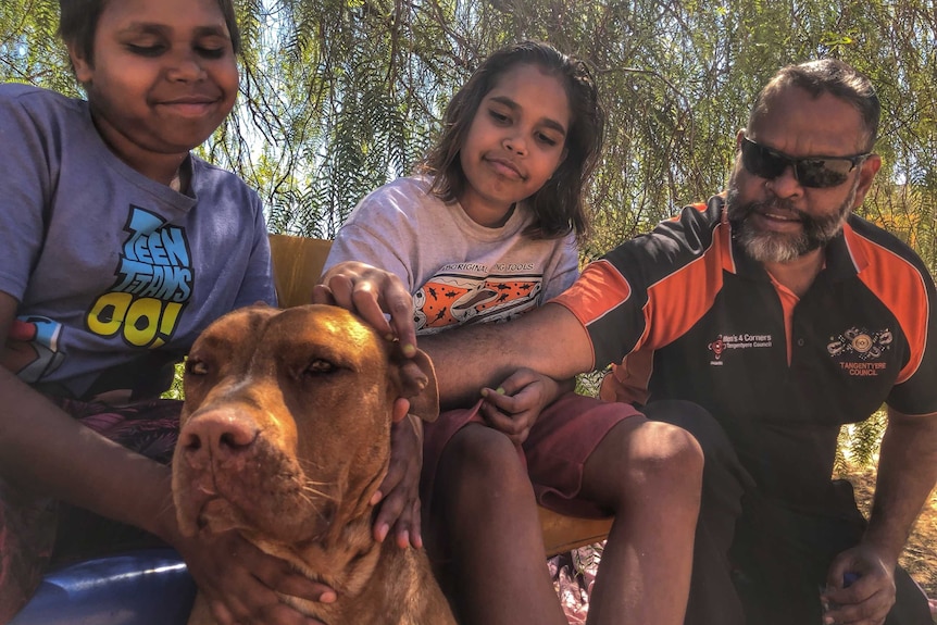 A man sits with his children and dog in a garden.