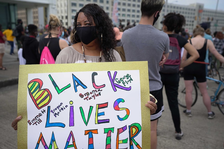 A black woman wearing a black mask and hoop earrings holds a sign saying Black Lives Matter with a crowd behind her