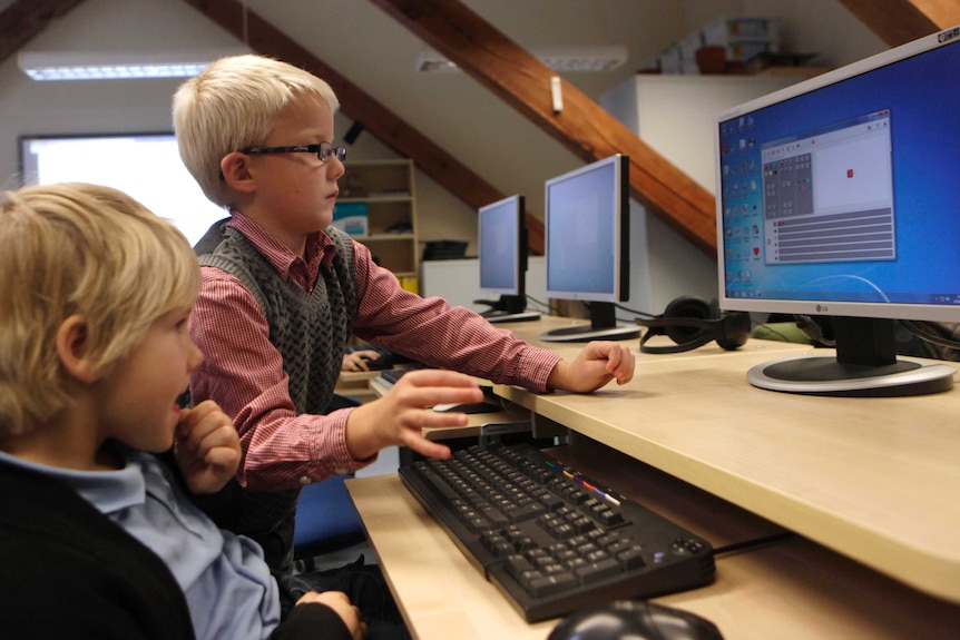 Two young children use computers in a classroom setting.