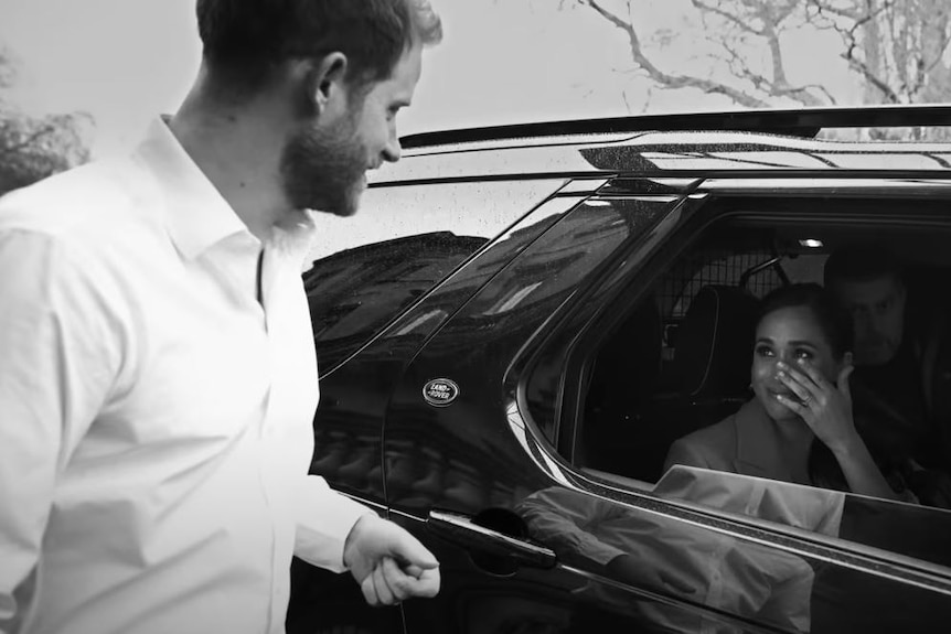 Prince Harry looks at Meghan through a car window as she brushes a tear from her face