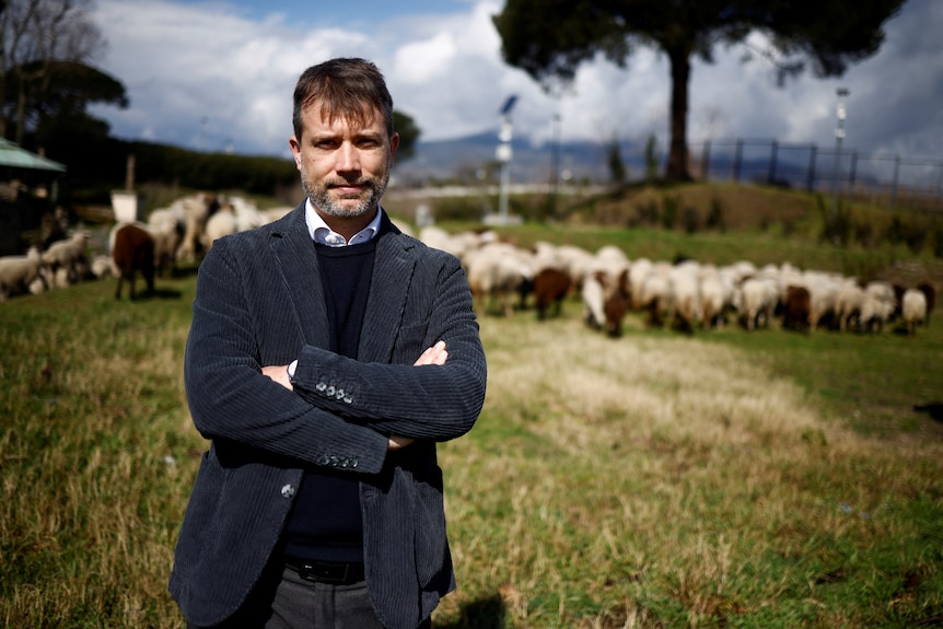 A young man in a suit stands with his arms folded in front of a flock of sheep.
