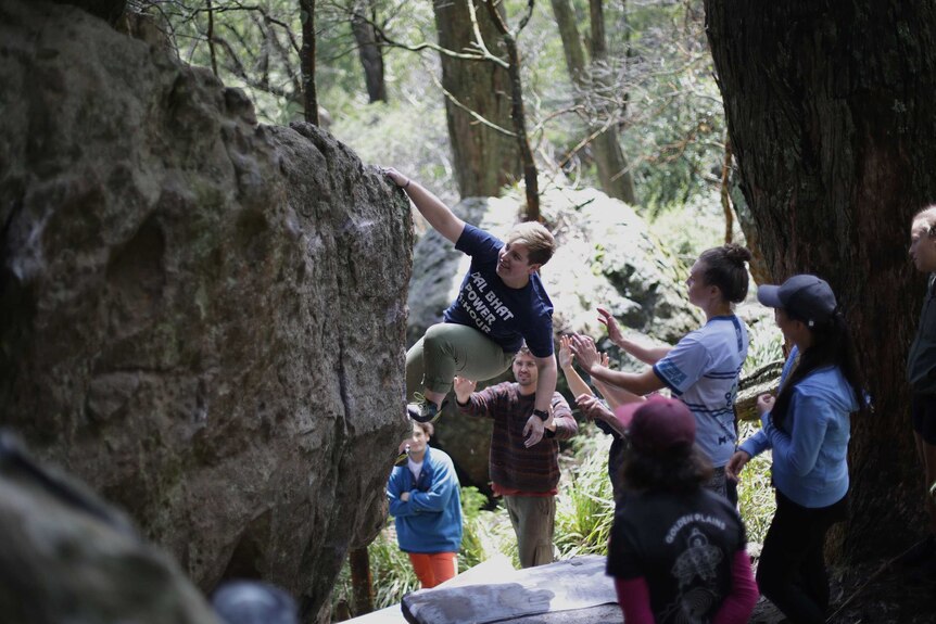A climber clambers up a rock as members of a team applaud and give support.
