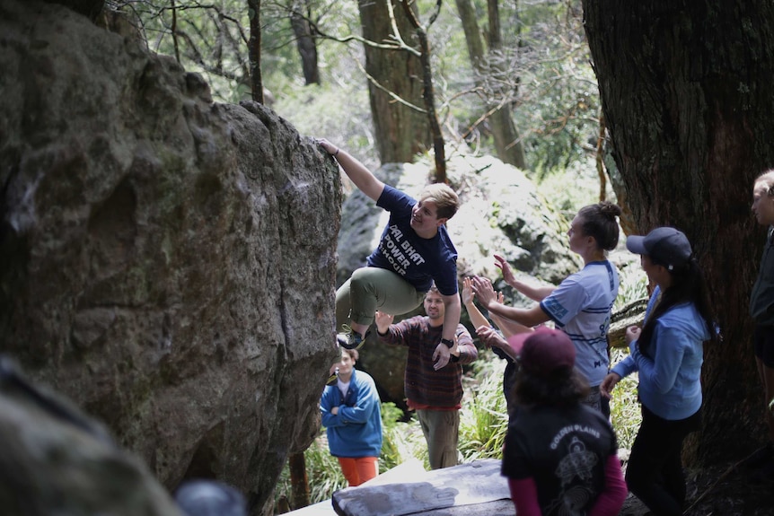 A climber clambers up a rock as members of a team applaud and give support.