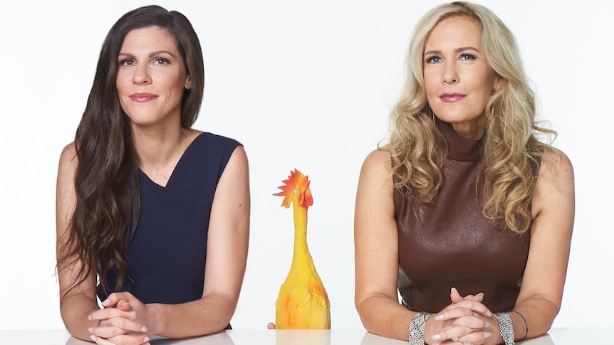 A brunette middle-aged woman and a blonde woman smirk while sitting behind a white desk. In between them is a rubber chicken.