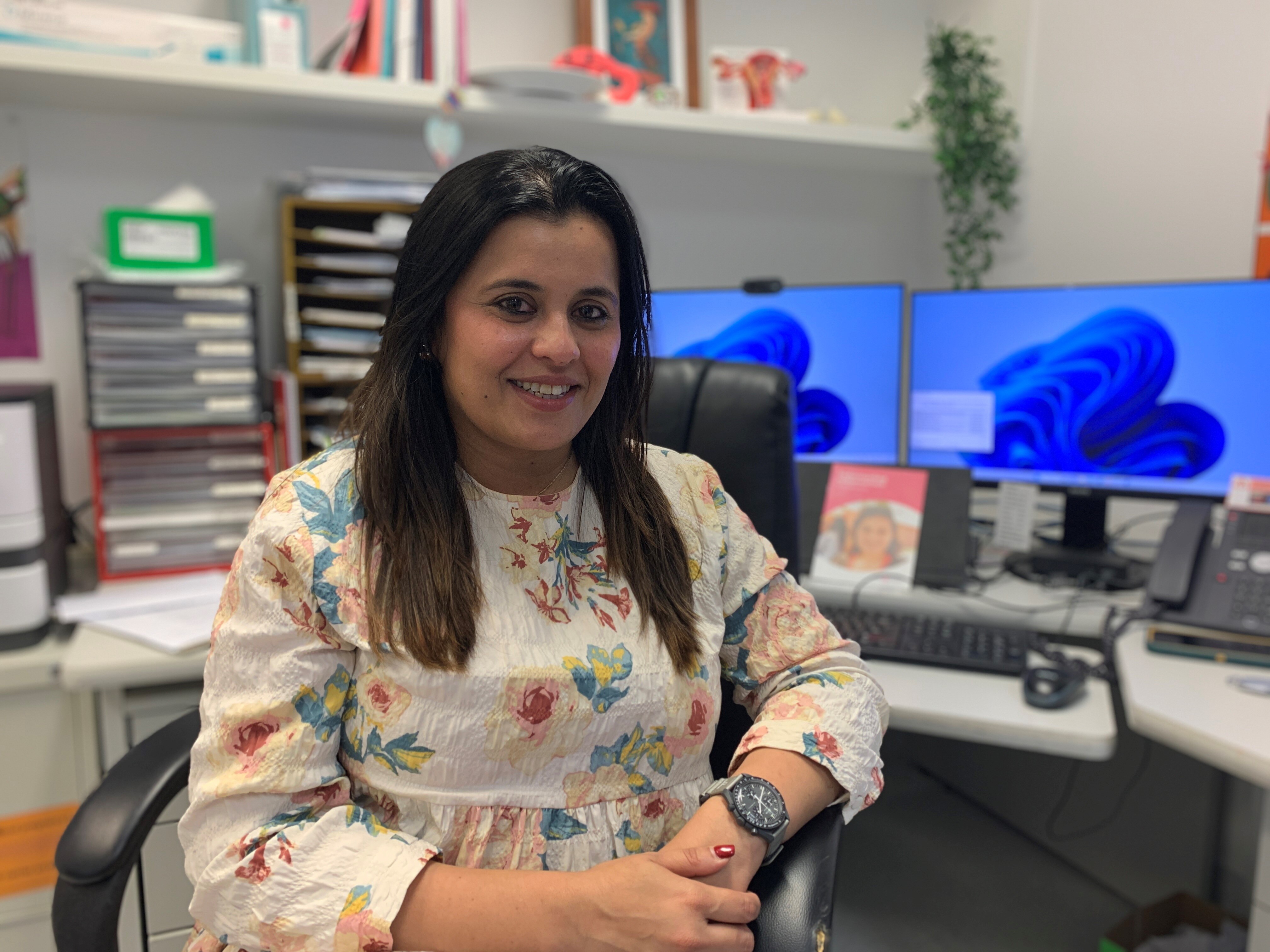 A woman with long dark hair, wearing a floral dress sitting at a desk with an endometriosis pamphlet in the background.