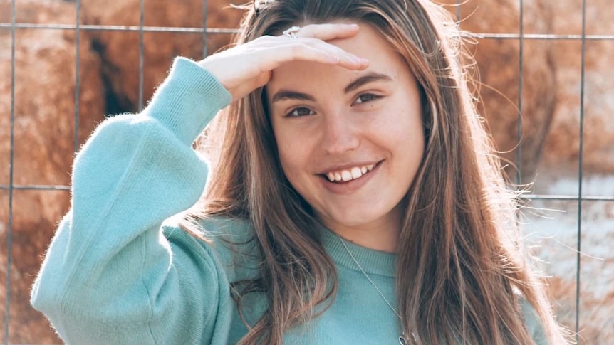 A young woman wearing a pale blue jumper holds her hand up to her forehead and smiles