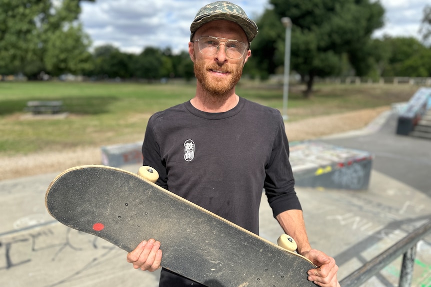 A man with a baseball cap and glasses holds a skateboard while looking at the camera